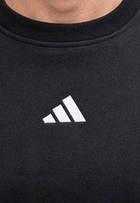 Λευκό λογότυπο Adidas με τρεις ρίγες κεντημένο στο στήθος ενός μαύρου μπλουζιού με στρογγυλή λαιμόκοψη που φοράει ένα άτομο.