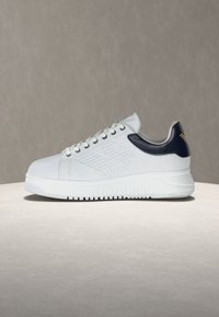 Λευκό αθλητικό παπούτσι χαμηλού ύψους με μαύρη ταμπελίτσα στη φτέρνα, ανάγλυφο λογότυπο φτερού στη πλευρά, λευκά κορδόνια και χοντρή σόλα πάνω σε ουδέτερη επιφάνεια και φόντο.