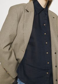 Blazer in tessuto a scacchi con tonalità beige e marrone, caratterizzato da spalle strutturate, rever a punta e tasche frontali. Indossato sopra una camicia a bottoni scura.