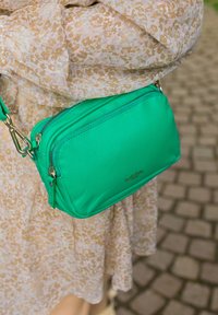 Zelená kožená crossbody taška s předními zipy, zlatými kovovými doplňky a hladkou texturou, nošena s šaty s květinovým vzorem.