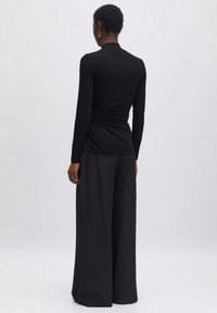 Schwarzes Langarmoberteil mit hochgeschlossenem Kragen, das an der Taille mit einem gerafften Design versehen ist. Kombiniert mit weit geschnittenen schwarzen Hosen aus glattem Material.