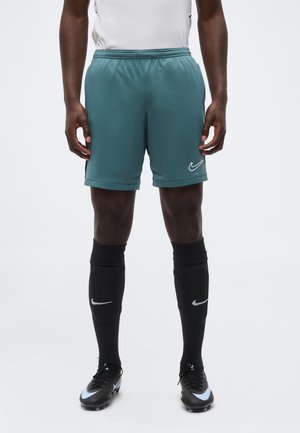 Atleta masculino con pantalones cortos verdes de Nike, calcetines negros hasta la rodilla de Nike y botas de fútbol negras con detalles azules, de pie sobre un fondo liso.