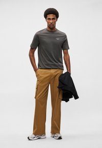 Pelēks īsām piedurknēm krekls, brūnas cargo bikses ar sānu kabatām un melna jaka rokās. Apavi: pelēki skriešanas apavi. Vienkāršs dizains, brīvā piegriezumā.