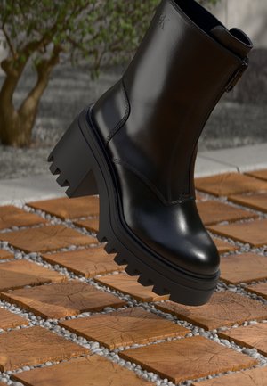 Black chunky heel ankle boot with zipper and textured wooden tiles on ground outdoors.