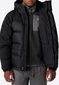 Sort dunjakke med hætte, polstret design, frontlynlås og velcro-detaljer. Båret over et gråt fleece-mellemlag. Grå bukser fuldender outfittet.