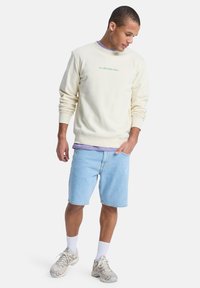 Sweat-shirt crème avec logo "Quiksilver" vert, porté sur une chemise lavande ; associé à un short en denim bleu clair et des baskets beige.