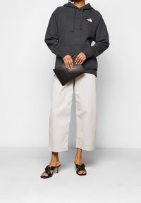 Felpa con cappuccio grigio scuro dotata di tasca frontale e logo, abbinata a pantaloni beige chiaro a gamba larga e sandali neri con tacco alto e dettaglio a nodo.