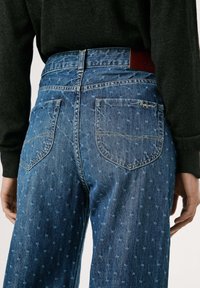Jean en denim avec un lavage bleu et un motif subtil, taille mi-haute, et deux poches arrière. Patch en cuir marron de la marque sur la ceinture.