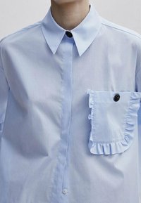 Chemise bleu clair boutonnée avec un col à pointes écartées, un détail de poche à volants, et un bouton noir décoratif sur le col et la poche.