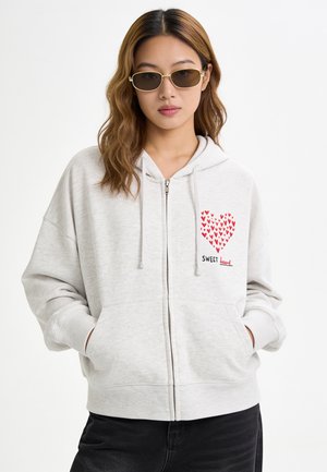 Jeune femme portant des lunettes de soleil rectangulaires et un sweat à capuche gris clair zippé avec un cœur rouge et le texte "Sweet heart" sur la poitrine.