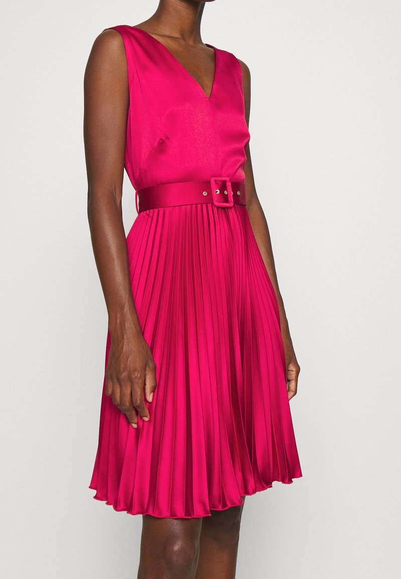 Robe fuchsia sans manches avec col en V, ceinture cintrée à la taille et jupe plissée longueur genou portée par une personne debout.