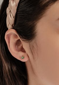 Goldene Ohrstecker mit einem grünen Edelsteindesign, das einem Blatt ähnelt, an das Ohr angelegt, mit sichtbarer, glatter Hauttextur.