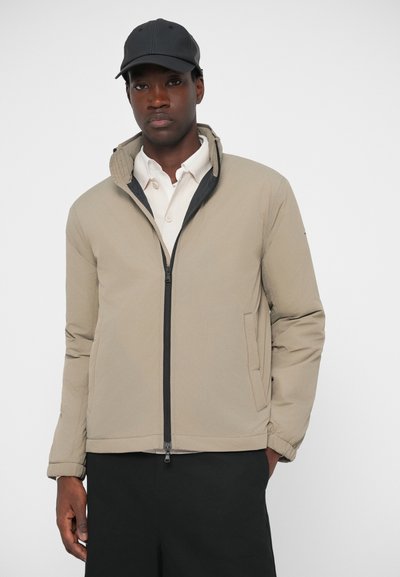 Jachetă bej căptușită cu fermoar negru, guler înalt și buzunare laterale, purtată cu o cămașă albă și o caschetă neagră. Textură netedă.