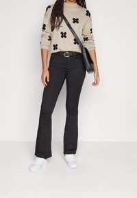 Beiger Pullover mit schwarzen Blumenmustern, kombiniert mit schwarzen Schlagjeans, einem schwarzen Gürtel und weißen Turnschuhen; eine Umhängetasche komplettiert den Look.