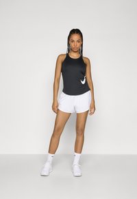 Zwarte mouwloze sporttop met het witte Nike-logo, gecombineerd met witte shorts en witte sneakers; gladde stof, casual pasvorm en minimalistisch ontwerp.