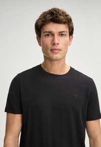 Zwarte katoenen t-shirt met een ronde hals, korte mouwen en een subtiele geborduurde detail op de linkerkant van de borst. Glad van textuur en aangesloten pasvorm.