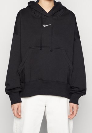 Person iført en sort Nike hættetrøje med en frontlomme og hvide joggingbukser, vist fra nakke til midt på låret, stående mod en ensfarvet baggrund.