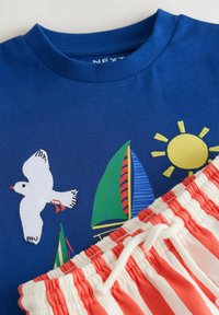 Chemise bleue avec mouette blanche brodée, voiliers colorés et soleil jaune, associée à un short rouge et blanc rayé avec un cordon blanc.