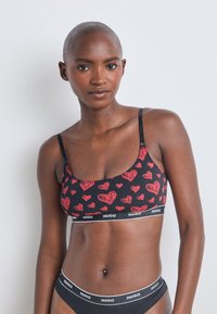 Svart bralette med rødt hjerteprint, justerbare stropper og en bred elastisk bånd med merkevarenavnet "HUGO" i hvitt.