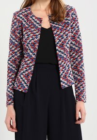 Blazer tissé en motif géométrique rouge, bleu et blanc. Encolure ronde, manches longues, associé à un top noir et un pantalon foncé.
