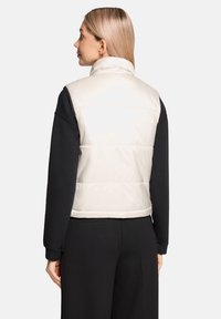 Pikowana kamizelka w jasnobeżowym kolorze, z wysokim kołnierzem, pikowany wzór, noszona na czarnej bluzce z długim rękawem, pokazana od tyłu. Gładka tekstura.