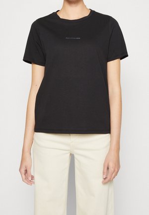 Personne portant un t-shirt noir à manches courtes avec un petit texte "Marc O’Polo Denim" et un pantalon beige clair sur un fond uni.