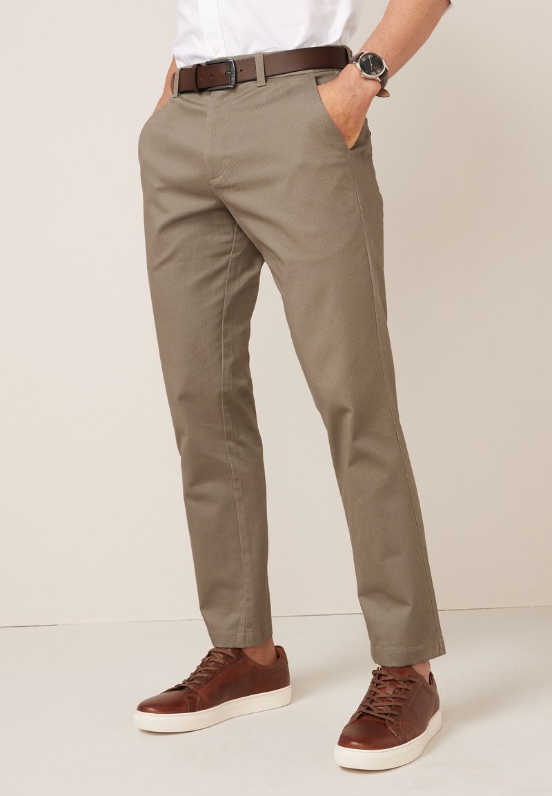 Beige slim-fit chino's met een gestructureerd oppervlak, zijzakken en een bruine leren riem, gecombineerd met bruine sneakers met witte zolen.