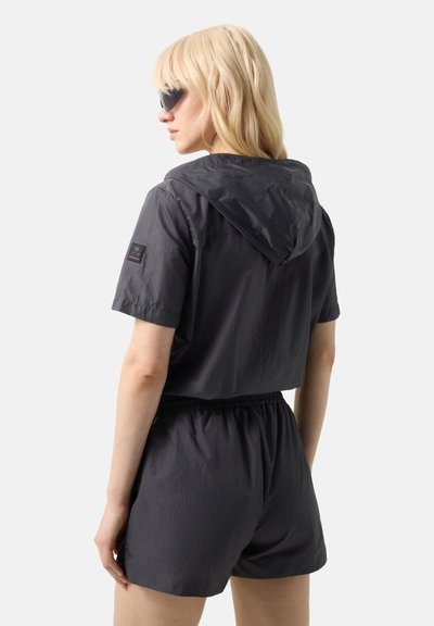 Lyshåret kvinde iført sort, kortærmet hættejakke og matchende shorts, set fra siden med sorte solbriller på.