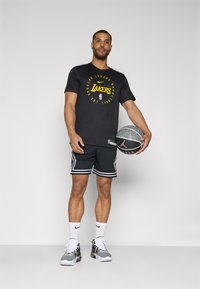 T-shirt noire avec le logo "Lakers" jaune, short noir avec bord blanc, chaussettes blanches et chaussures grises. Tient un ballon de basket avec une texture grise.