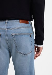 Svetlomodré rifľové nohavice s priamym strihom, s vreckami na zadnej strane, kontrastným šitím a hnedou koženou nášivkou s logom značky na opasku.