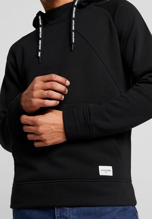 Mężczyzna w czarnej bluzie z kapturem z białą naszywką z logo Jack & Jones, trzymający lewe nadgarstek prawą ręką, na jednolitym szarym tle.