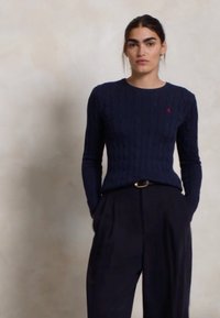 Suéter de punto trenzado en azul marino con mangas largas, cuello redondo y un pequeño logo; combinado con pantalones oscuros de talle alto que llevan un cinturón.