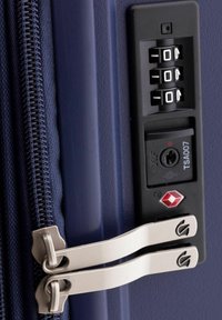 Dettaglio della chiusura lampo di una valigia blu navy con due tirazip in argento, una serratura TSA e un combinatore. La superficie è liscia, con una finitura esterna resistente.