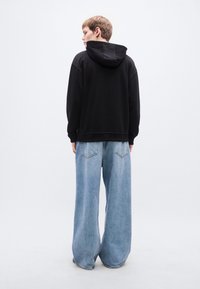 Μαύρο φούτερ με κουκούλα και άνετη γραμμή, σε συνδυασμό με ανοιχτόχρωμο μπλε τζιν παντελόνι με φαρδιά μπατζάκια και πίσω τσέπες. Λείες υφές, casual στυλ.