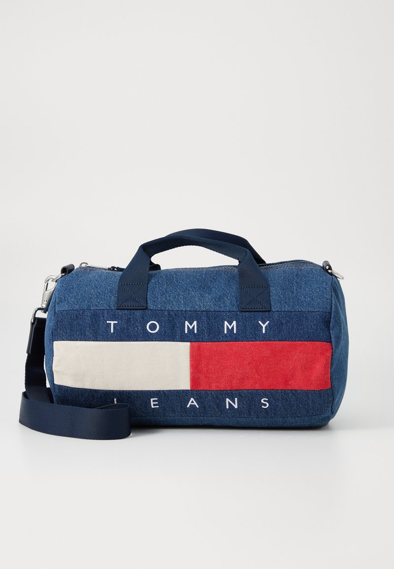 Sac de voyage en denim avec des accents rayés bleu marine, rouge et crème. Il dispose de deux poignées et d'une bandoulière réglable. Logo en texte "TOMMY JEANS."