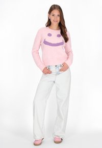 Rosa gestrickter Pullover mit einem lila Smiley-Gesicht-Design, kombiniert mit hellblauen weit geschnittenen Jeans und rosa Schuhen. Schlichte, weiche Textur.