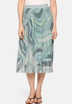 Femme portant une jupe midi plissée aux motifs abstraits verts, bleus et gris, associée à des sandales à anneau d'orteil marron sur un fond blanc.