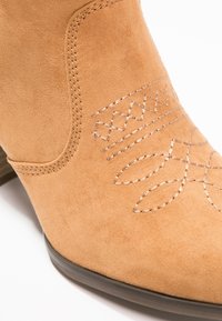 Bottine en daim beige avec couture décorative et talon bas marron foncé, présentée en gros plan sur fond blanc.