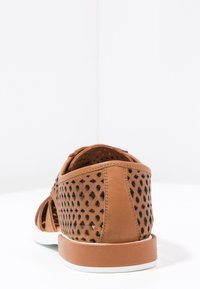 Scarpa in pelle marrone traforata con punta arrotondata, suola bianca e design strutturato. La parte posteriore presenta un pannello solido per supporto.