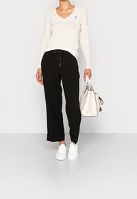 Pull-over crème en V, pantalon noir à cordon, et baskets blanches. Le modèle tient un sac rose pâle avec des accents noirs. Tenue simple et décontractée.