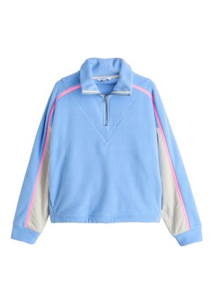 Person, die weiße Hose trägt, mit einer Hand in der Tasche und einer blauen Fleecejacke mit pinkem Streifen, eine blaue Kordel, die vom Bund hängt.