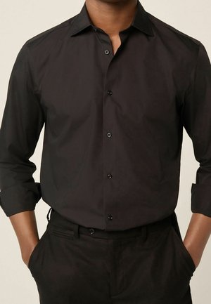 Mężczyzna ubrany w dopasowaną czarną koszulę na guziki z podwiniętymi rękawami i czarne spodnie, ręce w kieszeniach, neutralne tło.