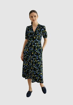 Mujer con vestido midi negro con estampado floral en azul y amarillo, mangas cortas abullonadas, escote en V y zapatos náuticos sin cordones sobre un fondo liso.
