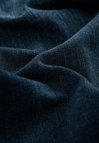 Tamno plavi denimski materijal s suptilnom tkanom teksturom, u dubokoj indigo boji s blagim sjajem. Površina pokazuje nabore i pregibe.