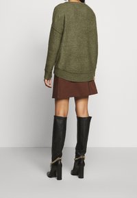 Pull oversize vert olive à col rond, associé à une jupe marron et des bottes noires montantes ornées d'un accent en chaîne métallique.