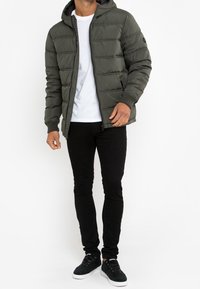 Padded oliwkowa kurtka z kapturem, zapinana na zamek, z prążkowanymi mankietami. Slim czarne spodnie i czarne sneakersy dopełniają zestaw.