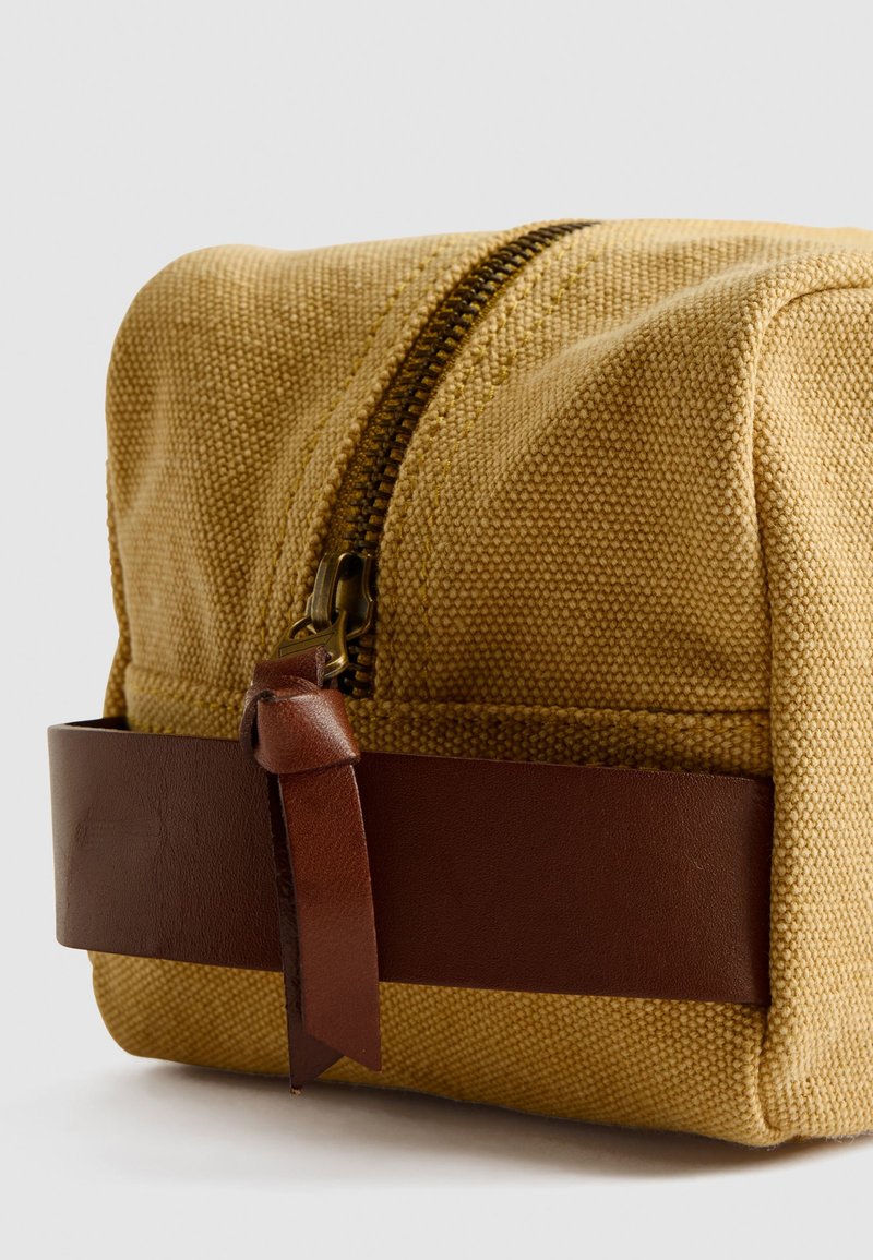 Borsetta da toiletta in tela color giallo senape, con una tracolla e zip in pelle marrone. La superficie ha una trama testurizzata per una migliore presa.