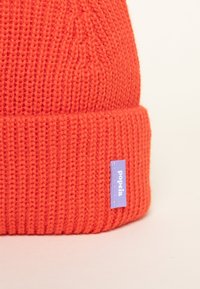Red knitted beanie with a folded brim, featuring a purple fabric tag labeled "popëa." The texture is thick and ribbed throughout.