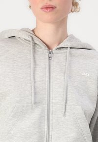 Sivi zip-up hoodie izrađen od mekog materijala, sa kapuljačom na vezanje, prednjim džepom i malim bijelim logotipom na prsima.