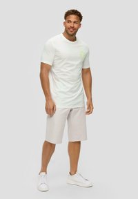 Witte T-shirt met korte mouwen met een lichtgroen cirkelvormig logo op de borst, gecombineerd met beige shorts tot de knie en witte sneakers.
